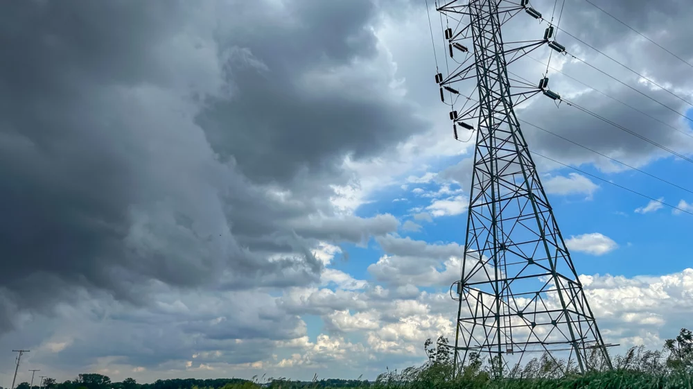 Transmission lines tower over green farmland alongside a rural road beneath a blue sky being overtaken by increasingly dark, gray clouds in the US Midwest.