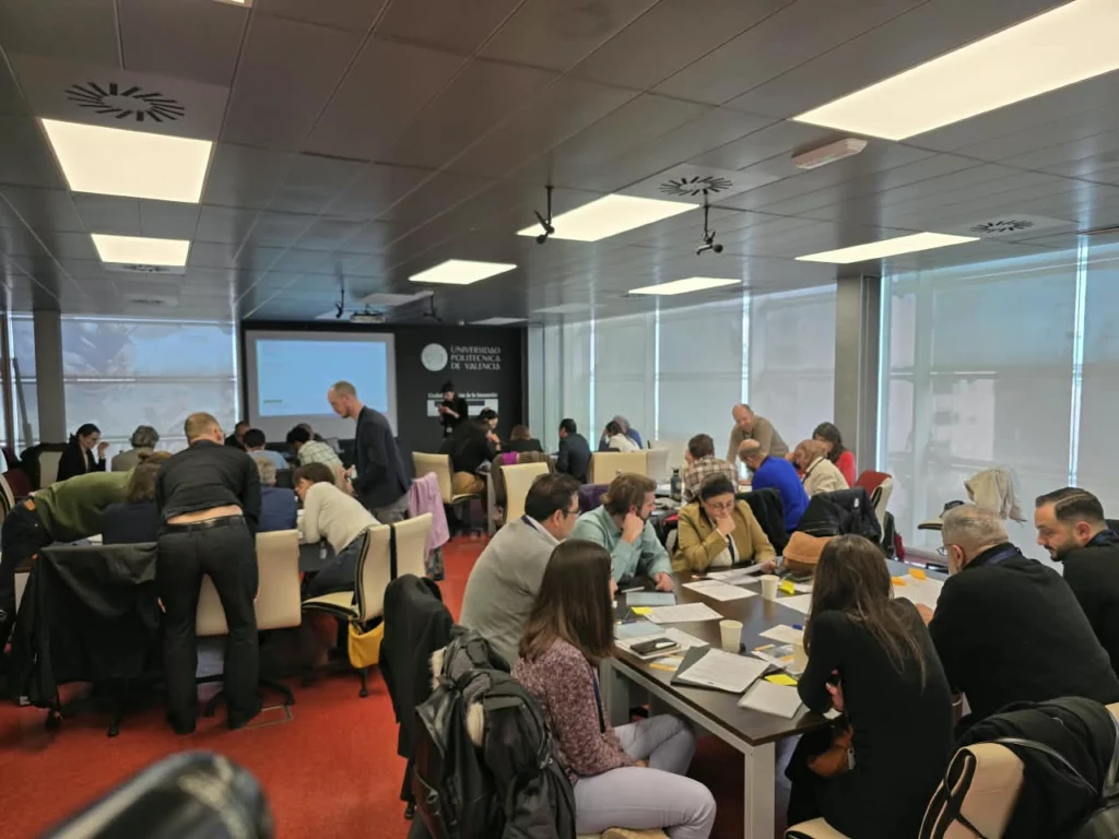 Photo shows a room of people sitting around tables, deep in thought and conversation, handling sheets of paper, tables covered in sheets of paper and paper cups.