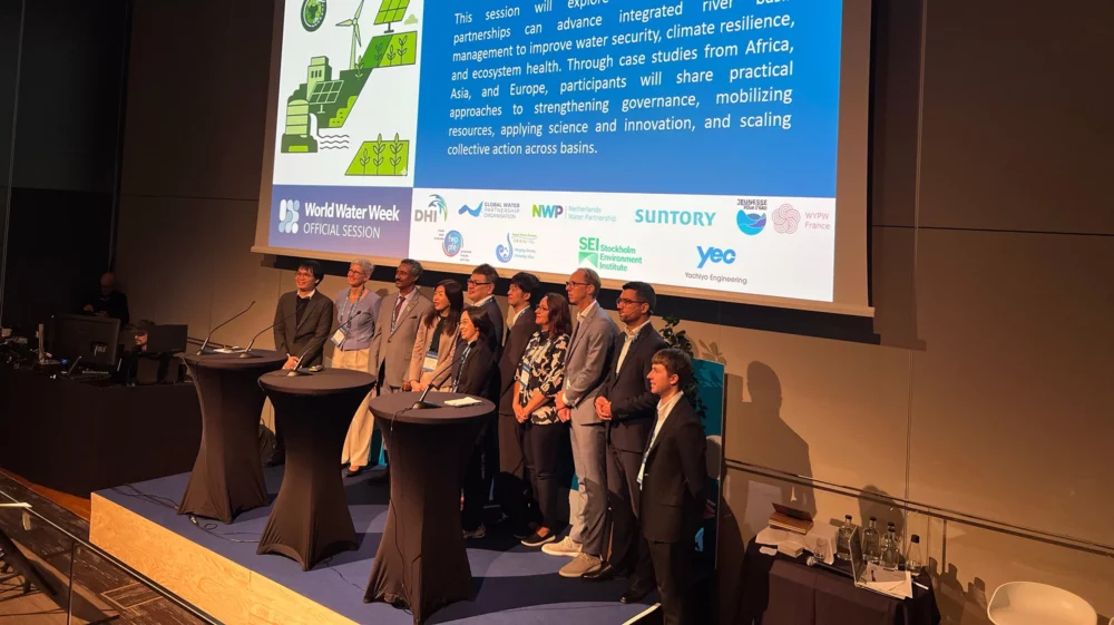 Eleven speakers stand together on stage facing the audience, below a screen showing the session title