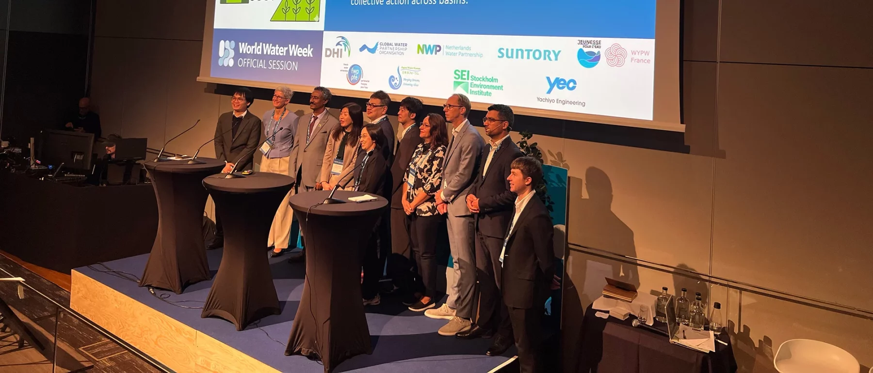 Eleven speakers stand together on stage facing the audience, below a screen showing the session title