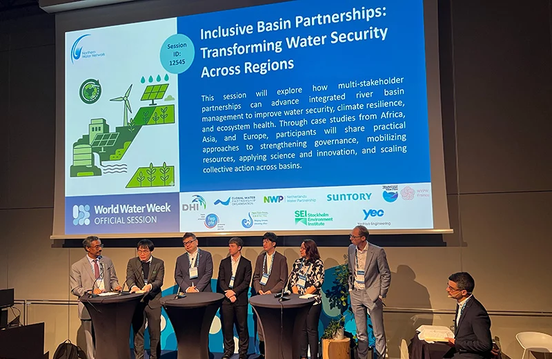 Eleven speakers stand together on stage facing the audience, below a screen showing the session title