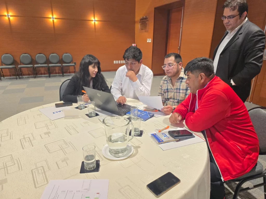 The image shows four people seated at a table around a laptop with a man behind them looking on.