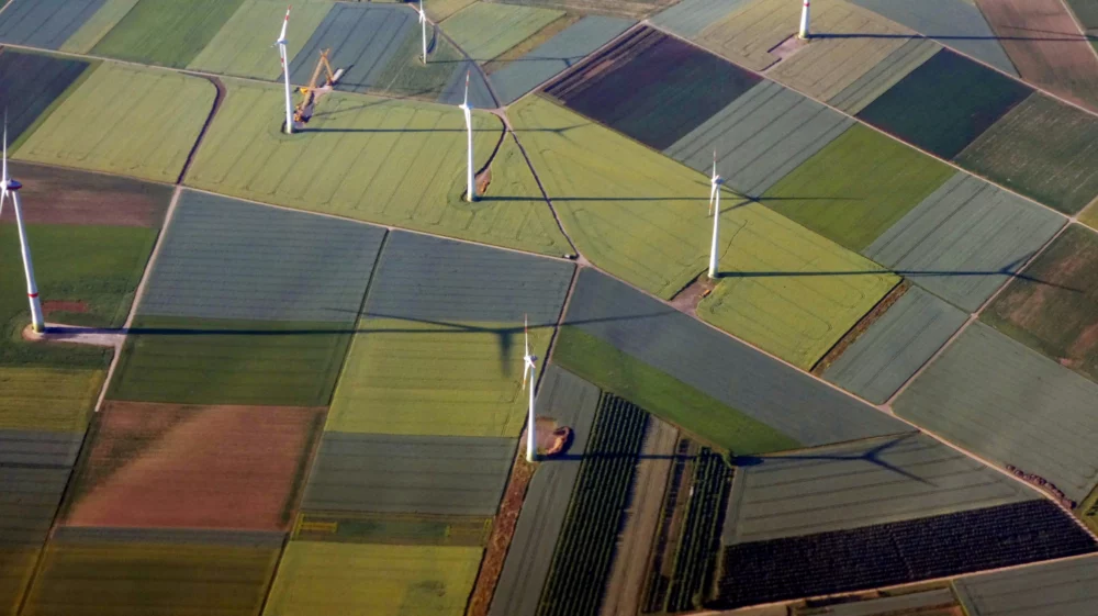 Aerial view of wind turbines casting shadows on a patchwork of different-colored agricultural fields in light green, dark green, rust, gray and charcoal.