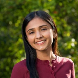 Ehina Srivastava smiles in a portrait against a green botanic background wearing a maroon button-up sweater