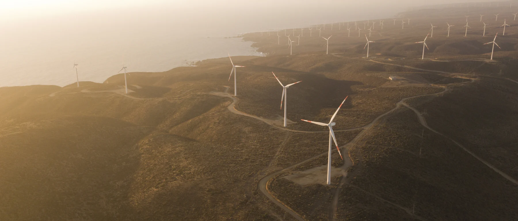 Aerial view of wind turbines dotting a rugged brown hillside along the coast in Chile at sunset.