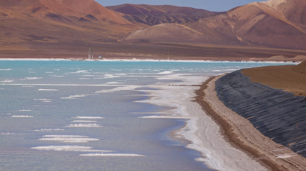 Landscape view of a lithium processing plant Susques, Argentina. It shows a pool of turquoise liquid where brine is extracted for 18 months against a dry, brown hilly horizon.