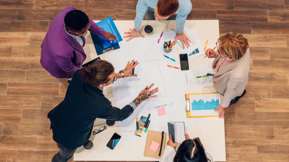 High angle shot of a diverse employees gathered in the office brainstorming while discussing new ideas for their new project. Multiracial coworkers gather in boardroom developing ideas at briefing.