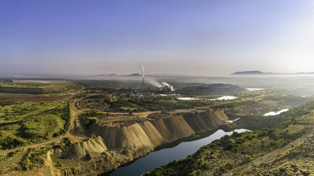 Aerial view of the Rhovan vanadium mining processing facility, seen on the horizon emitting plumes of smoke in a green and brown landscape beneath a cloudless sky, overlooking a river bluff
