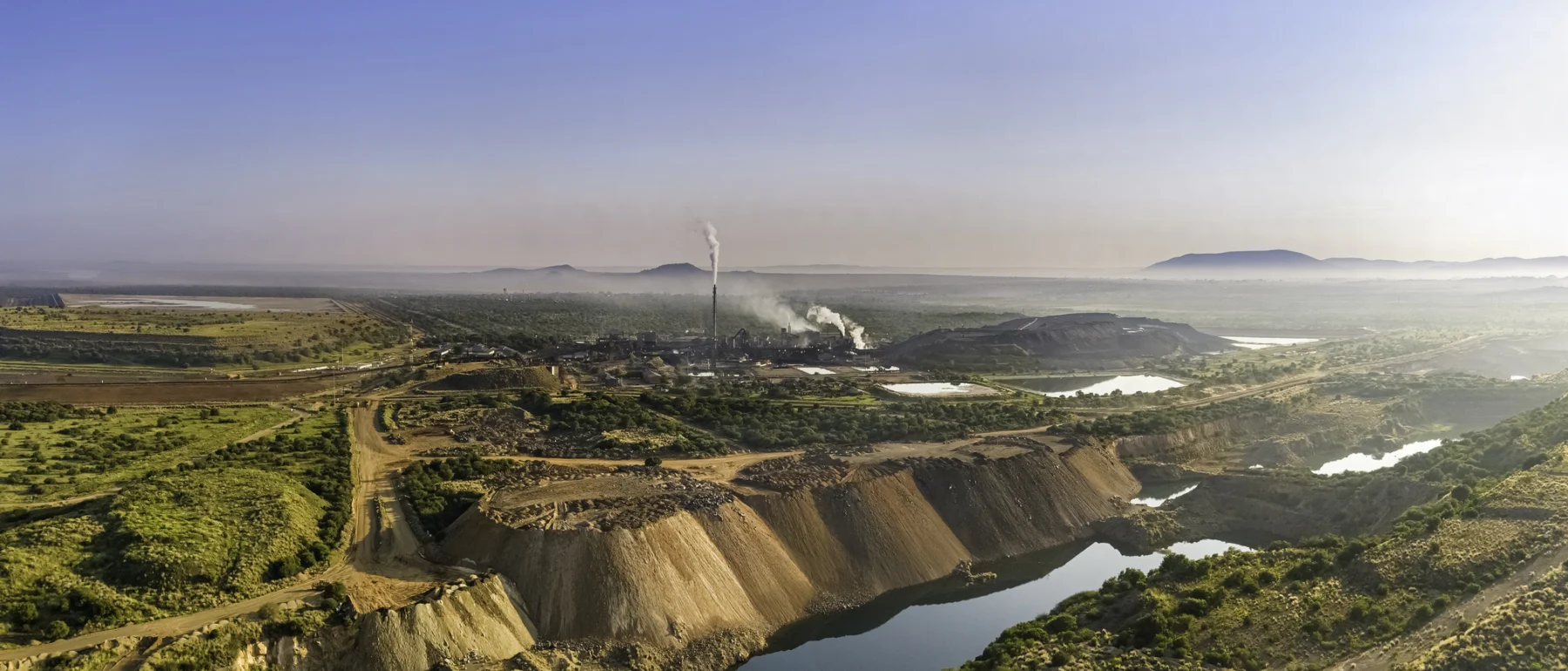 Aerial view of the Rhovan vanadium mining processing facility, seen on the horizon emitting plumes of smoke in a green and brown landscape beneath a cloudless sky, overlooking a river bluff