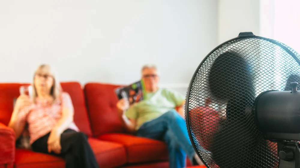 A senior couple sit on the sofa in front of an electric fan during a summer heat wave.