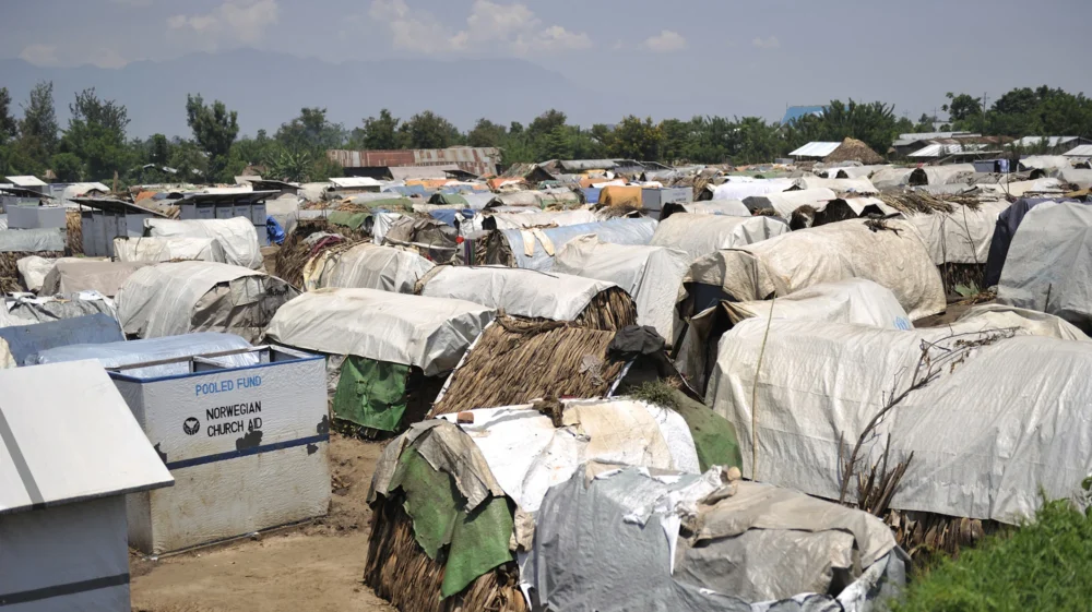 Shelters covered in white plastic tarps dominate the foreground to the horizon of trees, with a sign on a bin that says
