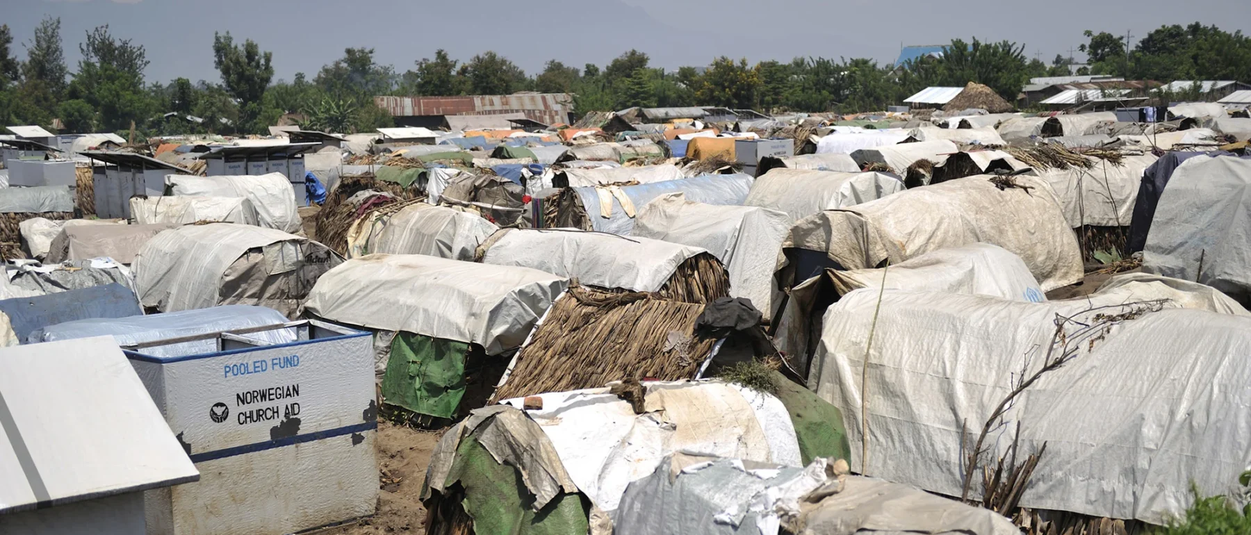 Shelters covered in white plastic tarps dominate the foreground to the horizon of trees, with a sign on a bin that says