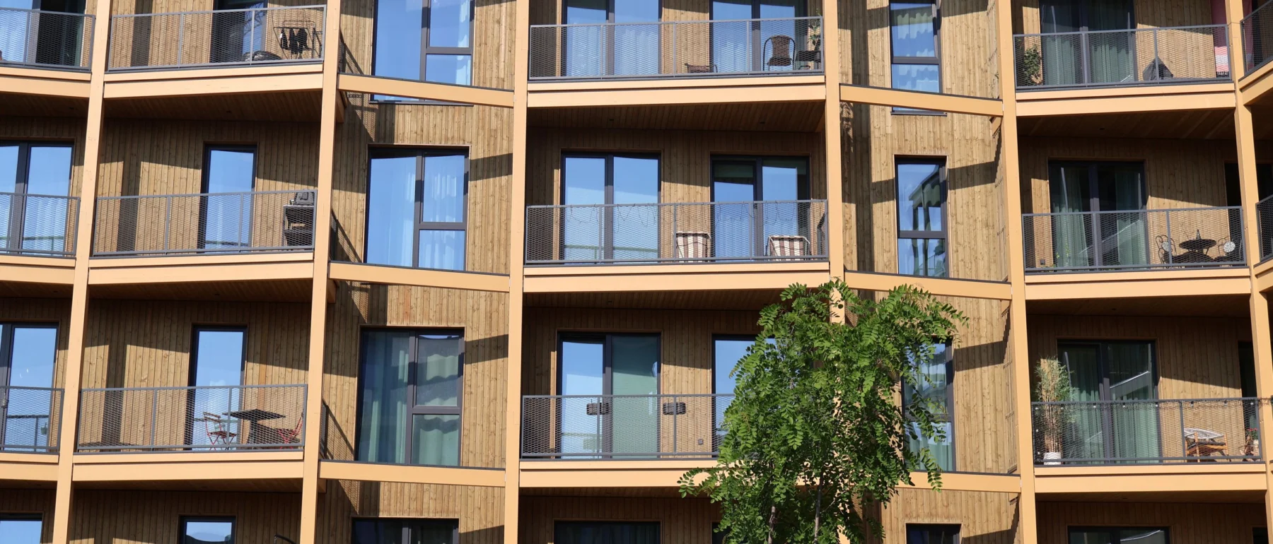 Modern timber-clad residential building with rows of balconies and a tree in the foreground