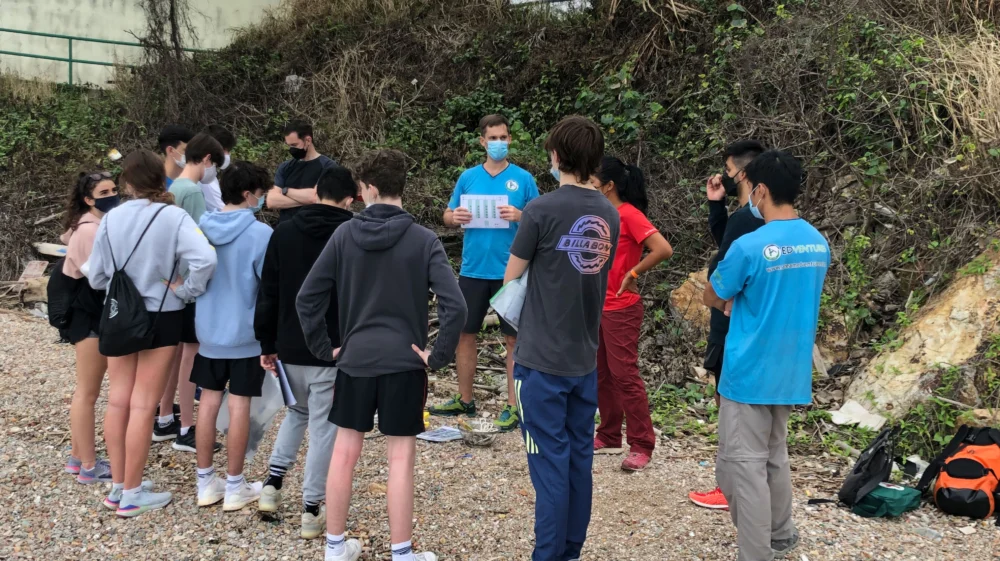 A group of teenage students stand on a pebble beach facing a faciliator. He is giving them instructions.