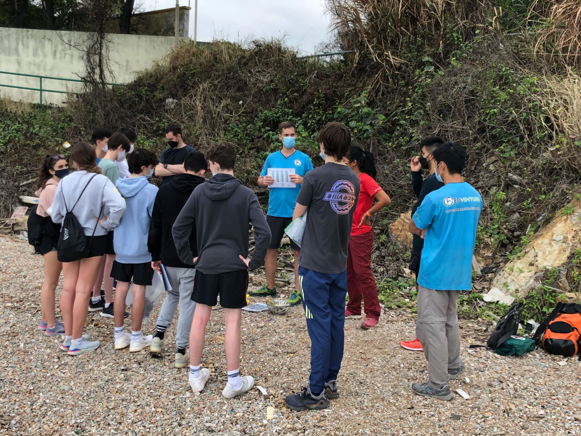 A group of teenage students stand on a pebble beach facing a faciliator. He is giving them instructions.