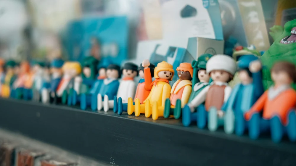 A group of little duplo people figurines have been lined up on a shelf. Most of the line is in soft focus; in the centre one smiling figure has been positioned with their hand raised, and is facing another, who is also smiling. Other toy figures in soft focus can be seen smiling too.