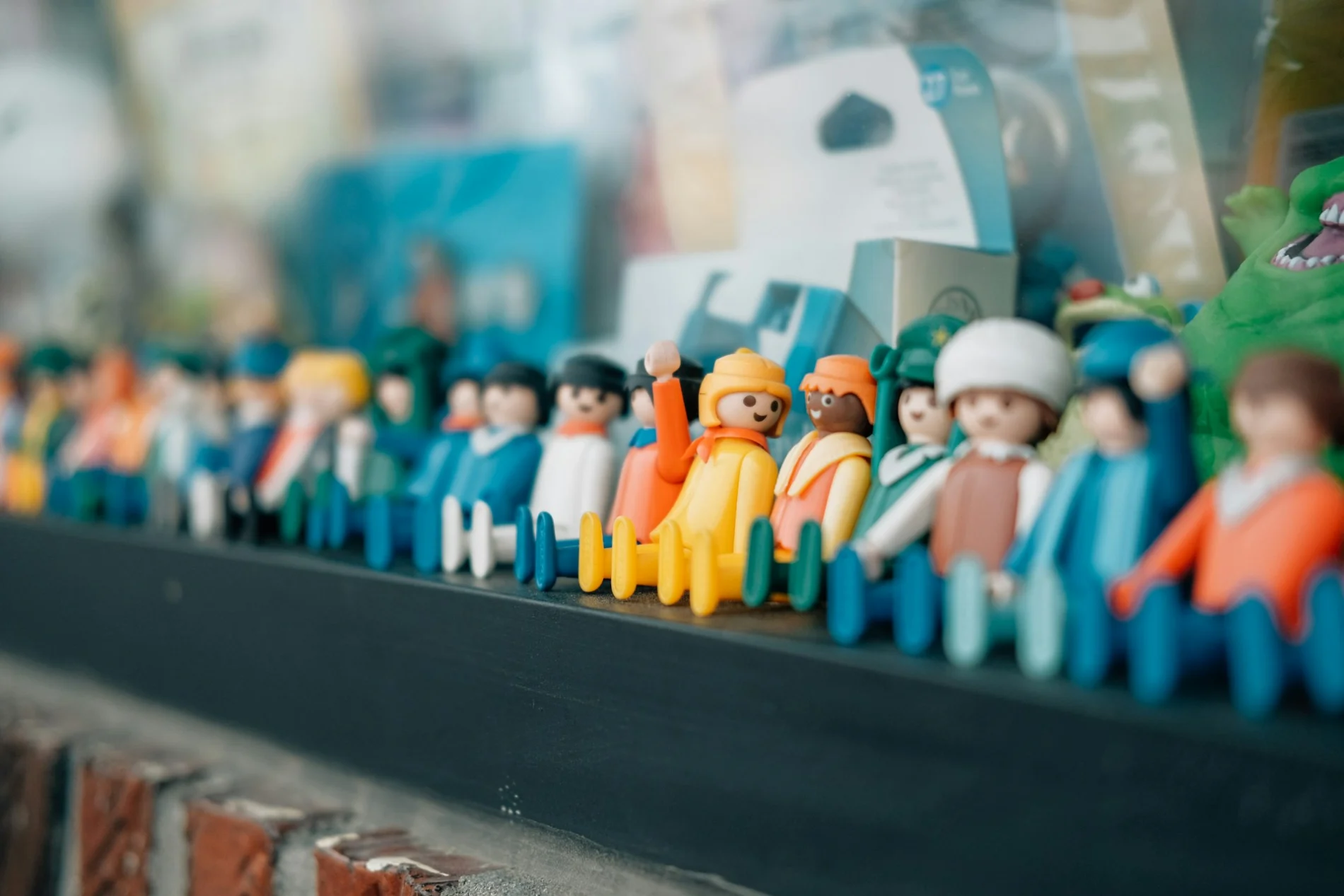 A group of little duplo people figurines have been lined up on a shelf. Most of the line is in soft focus; in the centre one smiling figure has been positioned with their hand raised, and is facing another, who is also smiling. Other toy figures in soft focus can be seen smiling too.