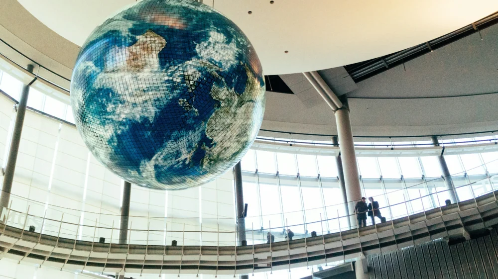 A large globe sculpture suspended in a circular glass atrium, with people standing on an upper-level walkway.