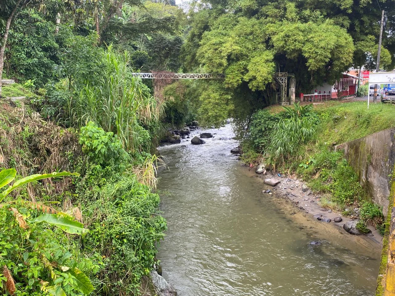 Campoalegre river flanked by green shores surrounded by trees and shrubs with man-made metal constructions, concrete reinforcements and a building and cars visible in the far background