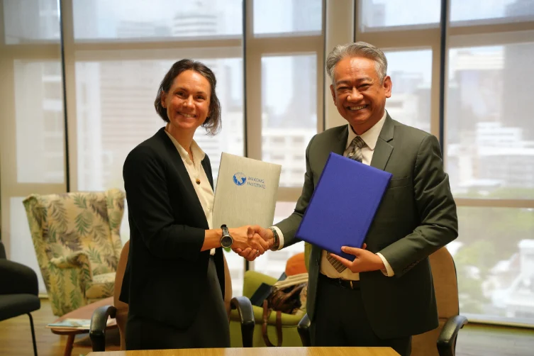 Marie Jürisoo shakes hands with Suriyan Vichitlekarn, each holding a signed memorandum of understanding folder, in an office with city views.