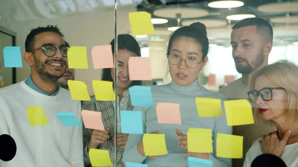 A group of people review post-in notes stuck to a large glass window. Their emotions vary, from smiles, to concentration.