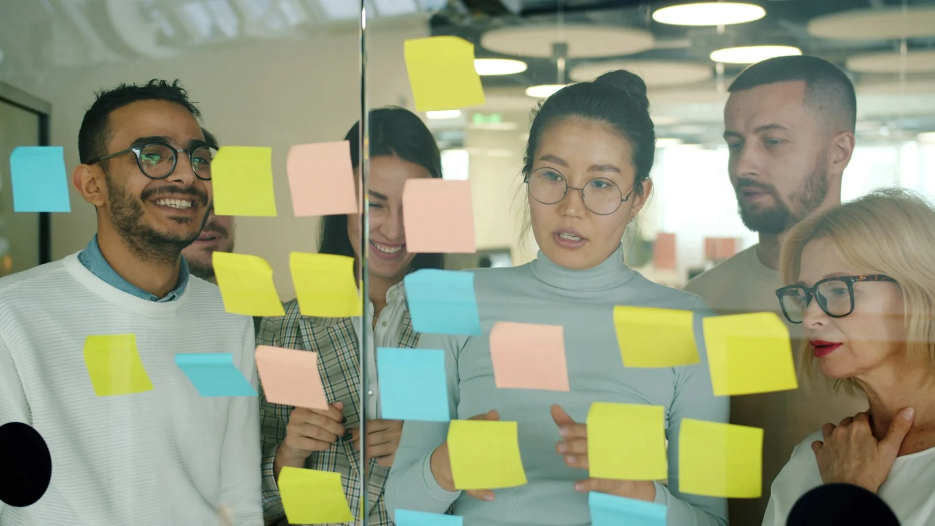 A group of people review post-in notes stuck to a large glass window. Their emotions vary, from smiles, to concentration.