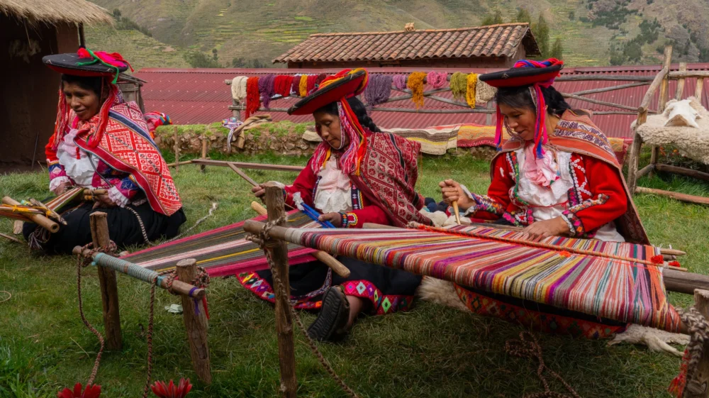 Three women weavers in colourful traditional local dress including festooned hats, weaving colourful alpaca wool, seated on the ground in Chumbe Community, Lamay, Sacred Valley, Peru.