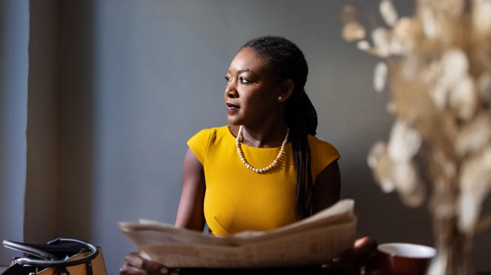 A woman in a yello top looks toward a window, with a newspaper in front of her, before a gray wall with beige leaves behind her.