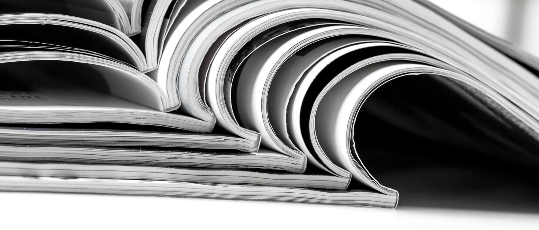 Eight magazines are stacked atop each other, open in the middle to create a line of Vs, with white page edges on a white background.
