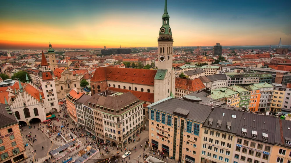Aerial view of Munich, Germany city skyline against a pink and blue twilight sky, with pedestrians filling the city square below.