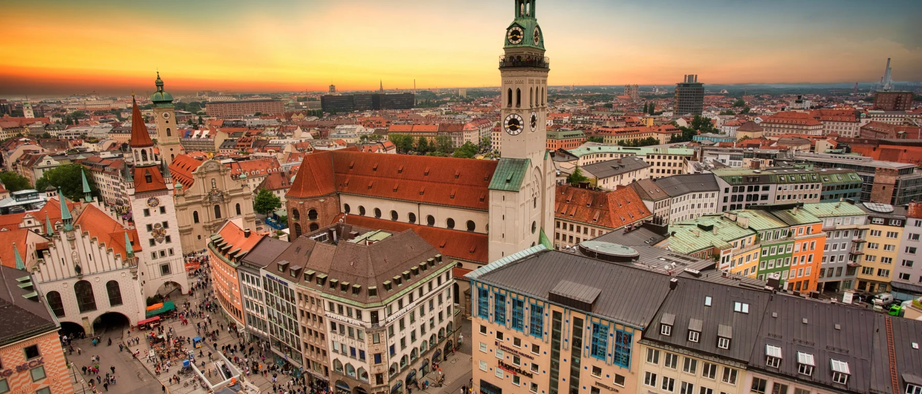 Aerial view of Munich, Germany city skyline against a pink and blue twilight sky, with pedestrians filling the city square below.