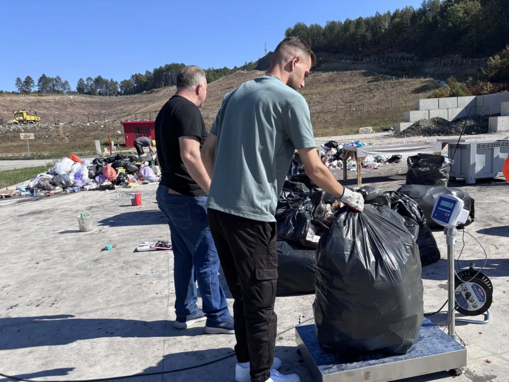 Two men weigh a full bag of sorted waste on a platform scale at a landfill sorting area, while others work nearby.