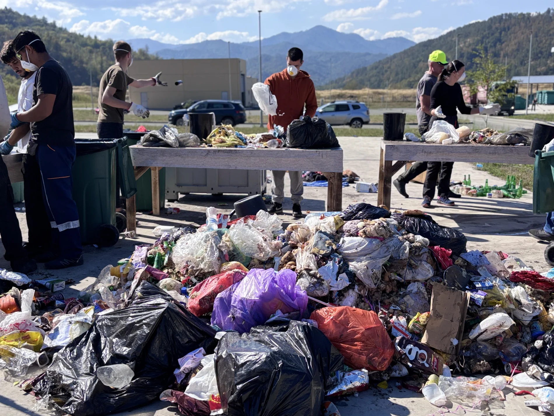 People in face masks and gloves sort mixed household waste at outdoor tables during a waste composition study. A large pile of bagged and loose rubbish sits in the foreground, with mountains behind.