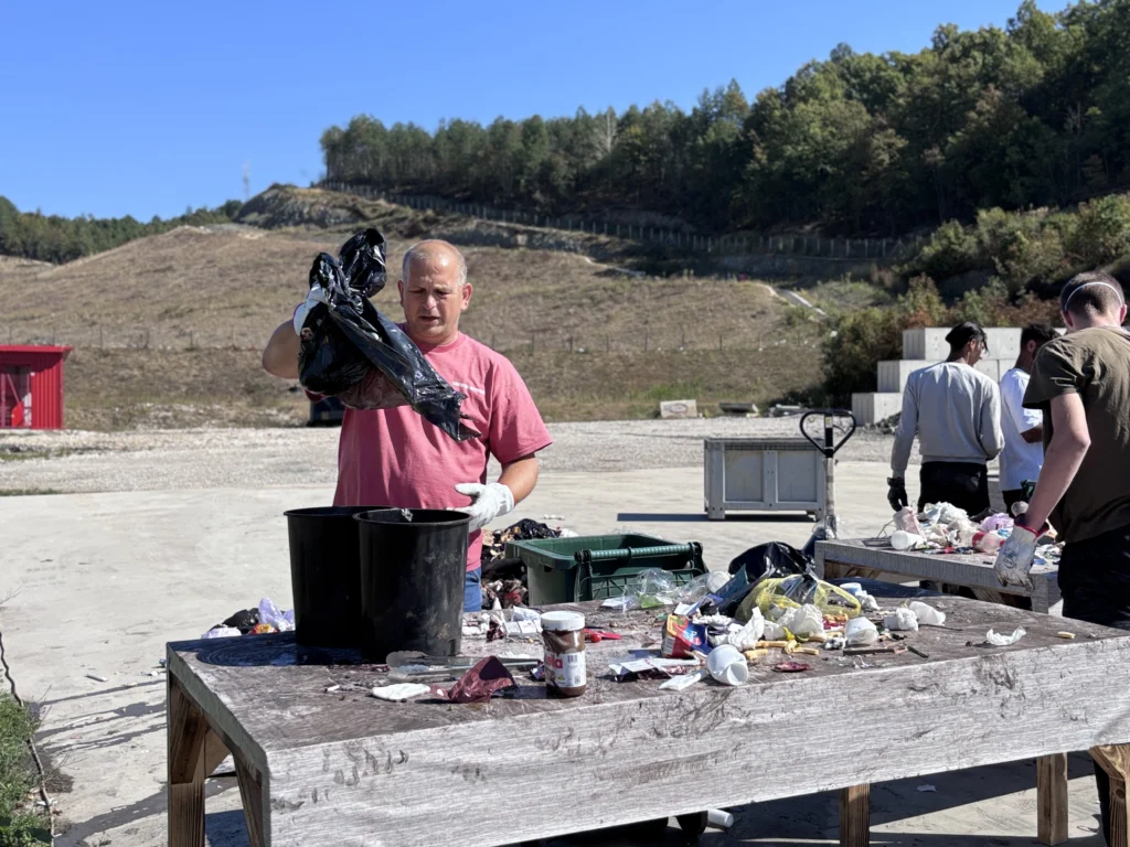 A man with red t-shirt sorting waste in an outdoor space behind the table full of waste.