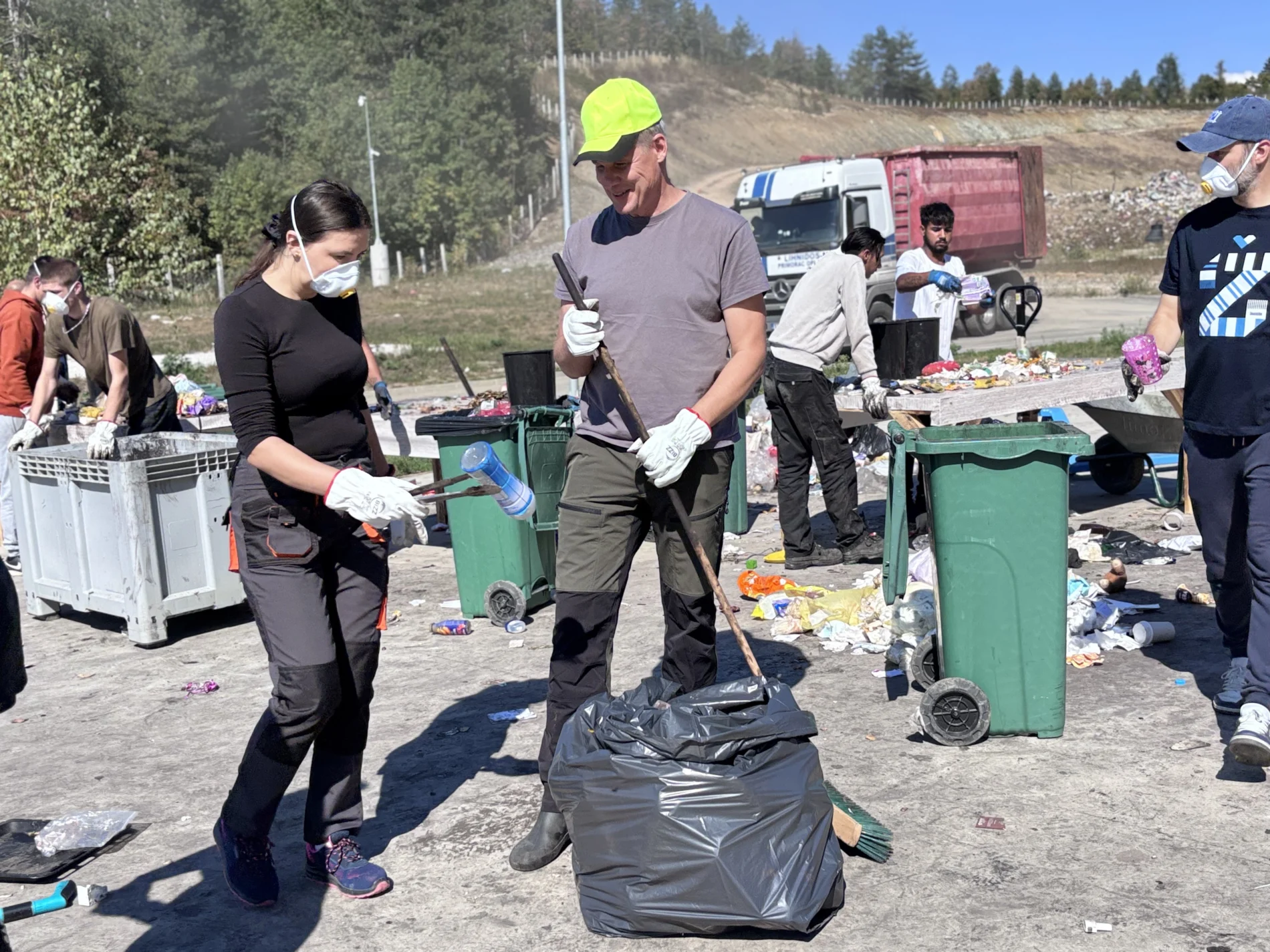 A woman holds up a plastic bottle while a man presses waste into a bin bag with a stick. Behind them, a group in masks and gloves sorts waste at tables and into wheelie bins.