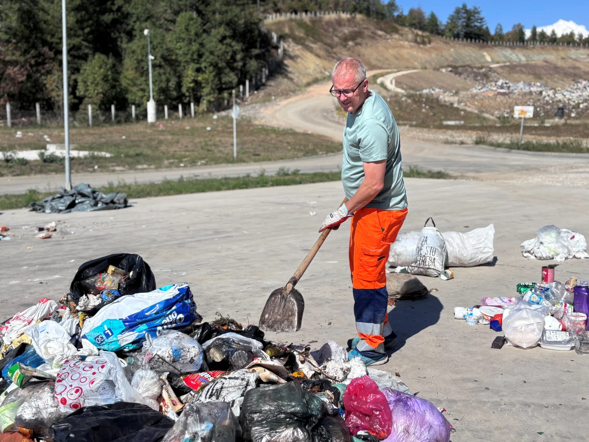 A man in high-visibility work trousers and gloves shovels through a pile of mixed household waste on a concrete sorting area. Sorted plastic packaging lies to one side, and a landfill slope rises in the background.