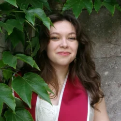 Katia wears a white dress and crimson graduation stole while peering out from behind vertical trails of ivy, with brown textured hair falling on her shoulders