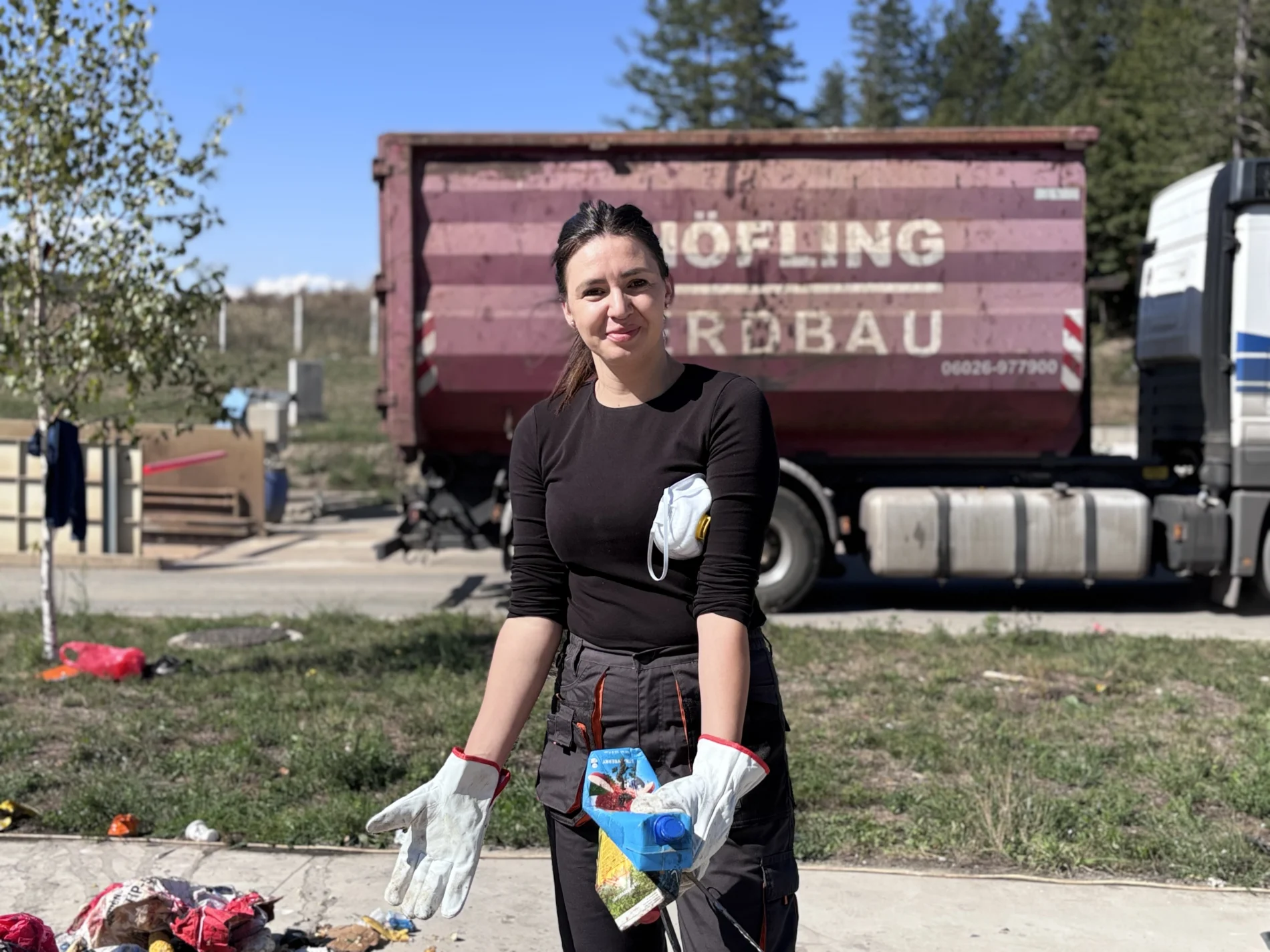 A woman in work gloves and workwear holds sorted packaging waste, including a drinks carton, at a waste sorting site. A respirator mask hangs from her arm, and a waste container truck is parked behind her.