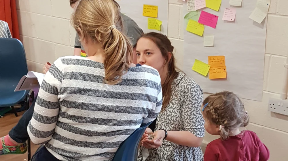A woman crouches on the floor in front of a wall covered in colourful sticky notes. She is talking to a woman sitting on a chair and a toddler, both of whom have their back to the camera.