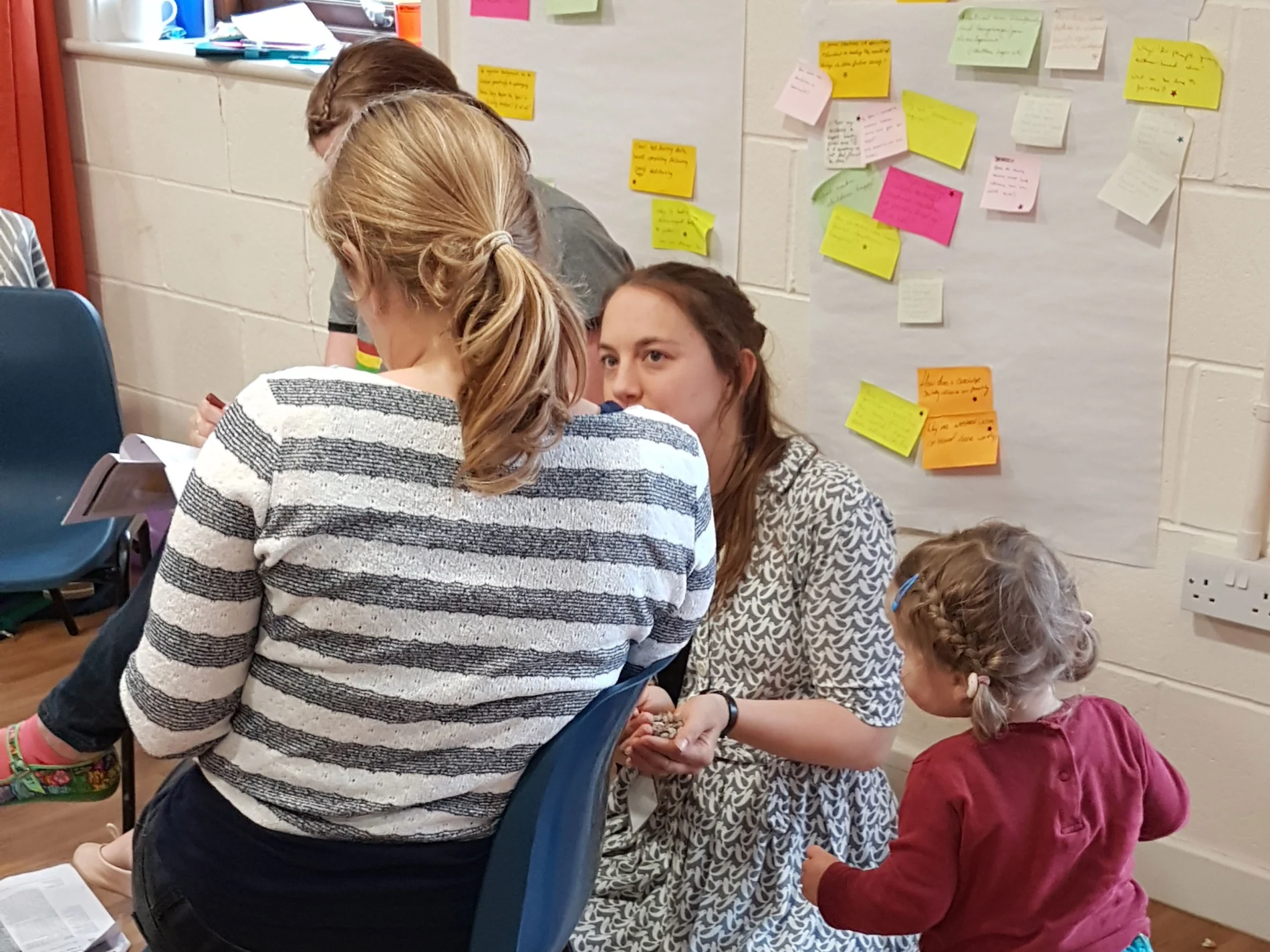 A woman crouches on the floor in front of a wall covered in colourful sticky notes. She is talking to a woman sitting on a chair and a toddler, both of whom have their back to the camera.