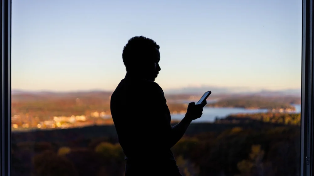 A person silhouetted against a scenic view, using a smartphone by the window at twilight.