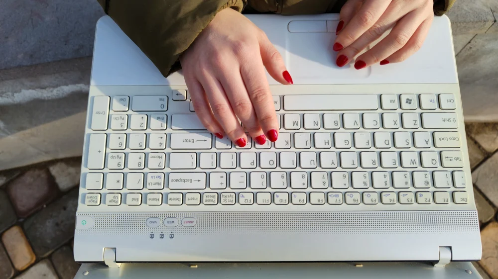 Aerial view of a woman's hands with red-varnished nails on a laptop keyboard. The woman is outside and appears to be sitting on something concrete on stone-paved ground.