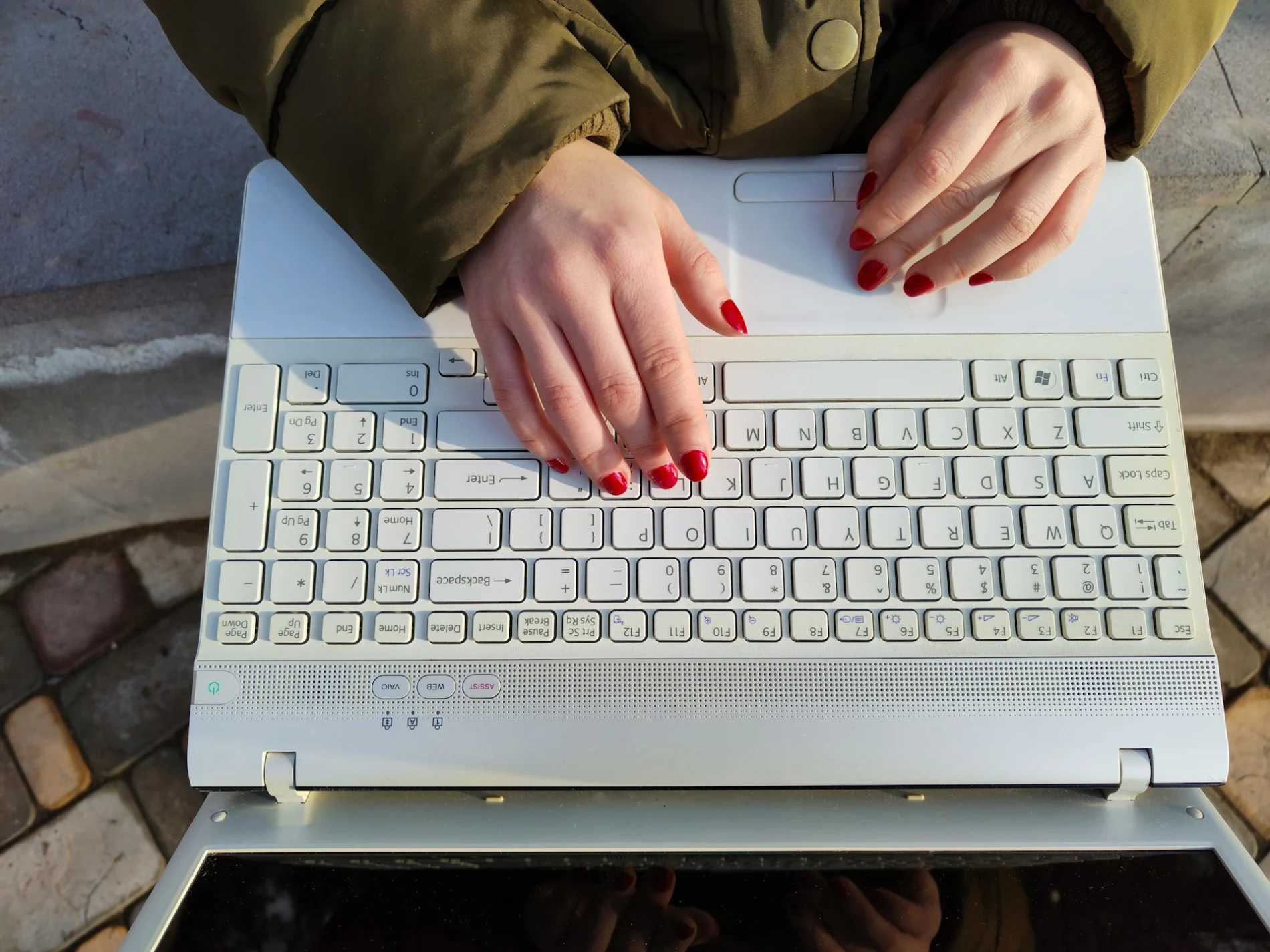 Aerial view of a woman's hands with red-varnished nails on a laptop keyboard. The woman is outside and appears to be sitting on something concrete on stone-paved ground.